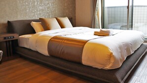 Una cama grande con estructura de madera y ropa de cama blanca, una mesita de noche con lámpara, y vista de la ciudad a través de una ventana.