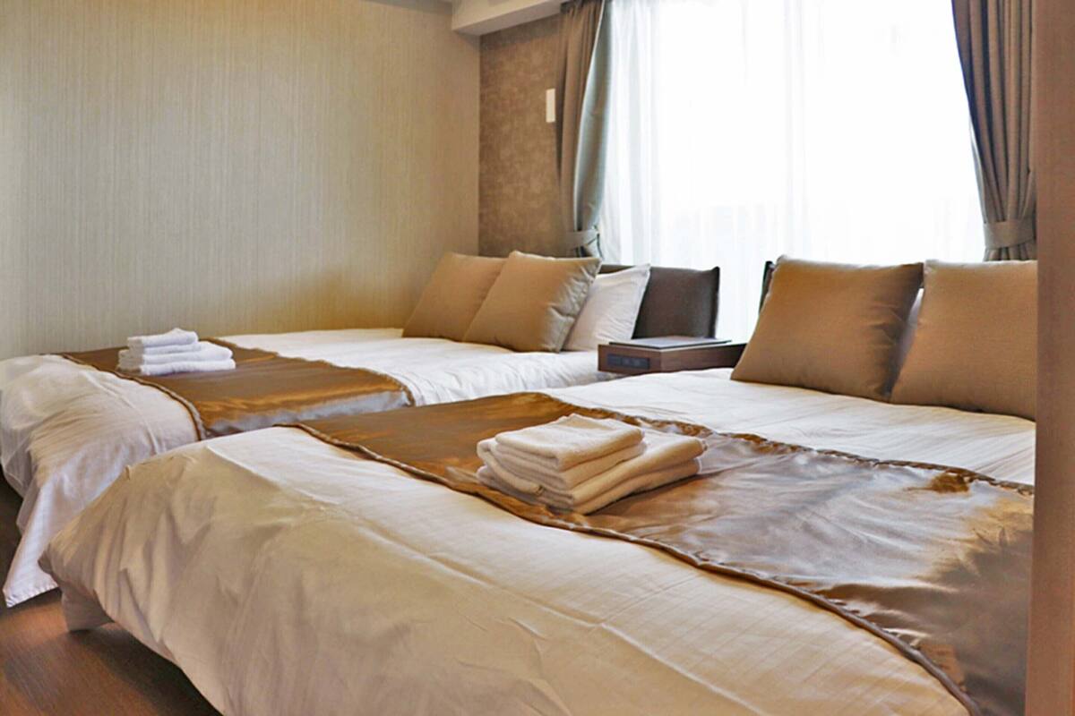 Une chambre d’hôtel avec deux lits, chacun doté d’une tête de lit marron, de draps blancs et d’un couvre-lit marron qui ressemble à du cuir. Une table de chevet se trouve entre les deux lits, avec une lampe et une petite table.