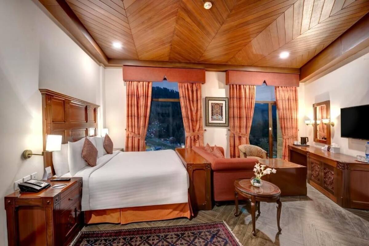 A hotel room with a wooden ceiling, a bed with white linens, a sofa, a small table with a vase, and a TV on a wooden cabinet.
