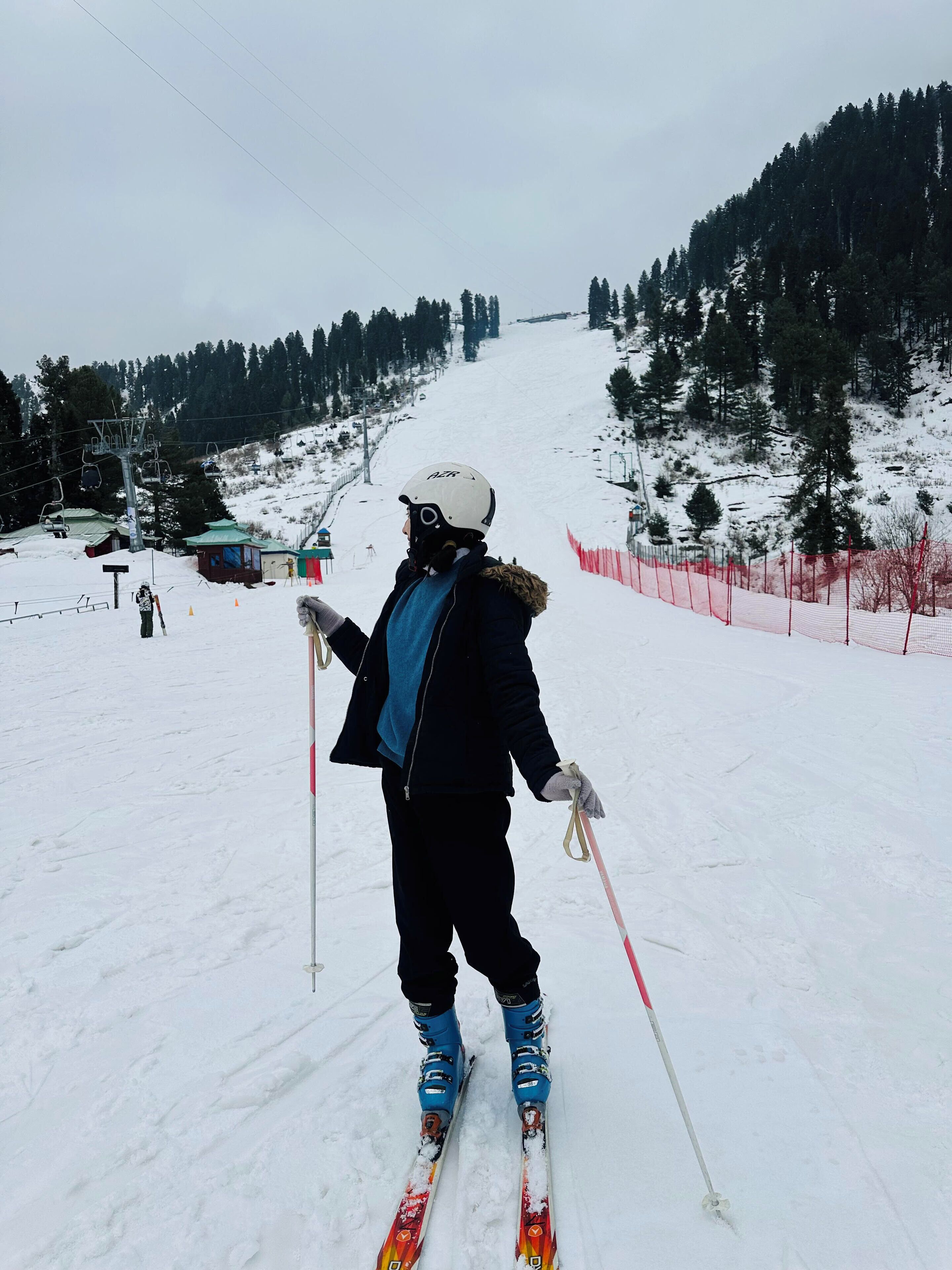 Una persona esquía por una pendiente nevada, con bastones de esquí, rodeada de árboles y un telesilla al fondo.