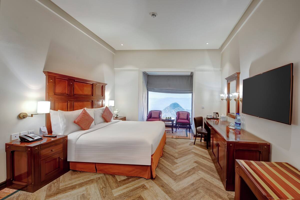 A hotel room with a large bed, wooden furniture, a flat-screen TV, and a view of mountains through a window.