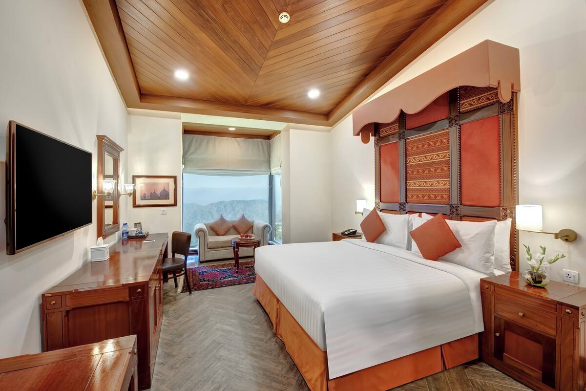 A hotel room with a large bed, wooden ceiling, a desk with a flat-screen TV, a sofa, and a view of mountains through the window.