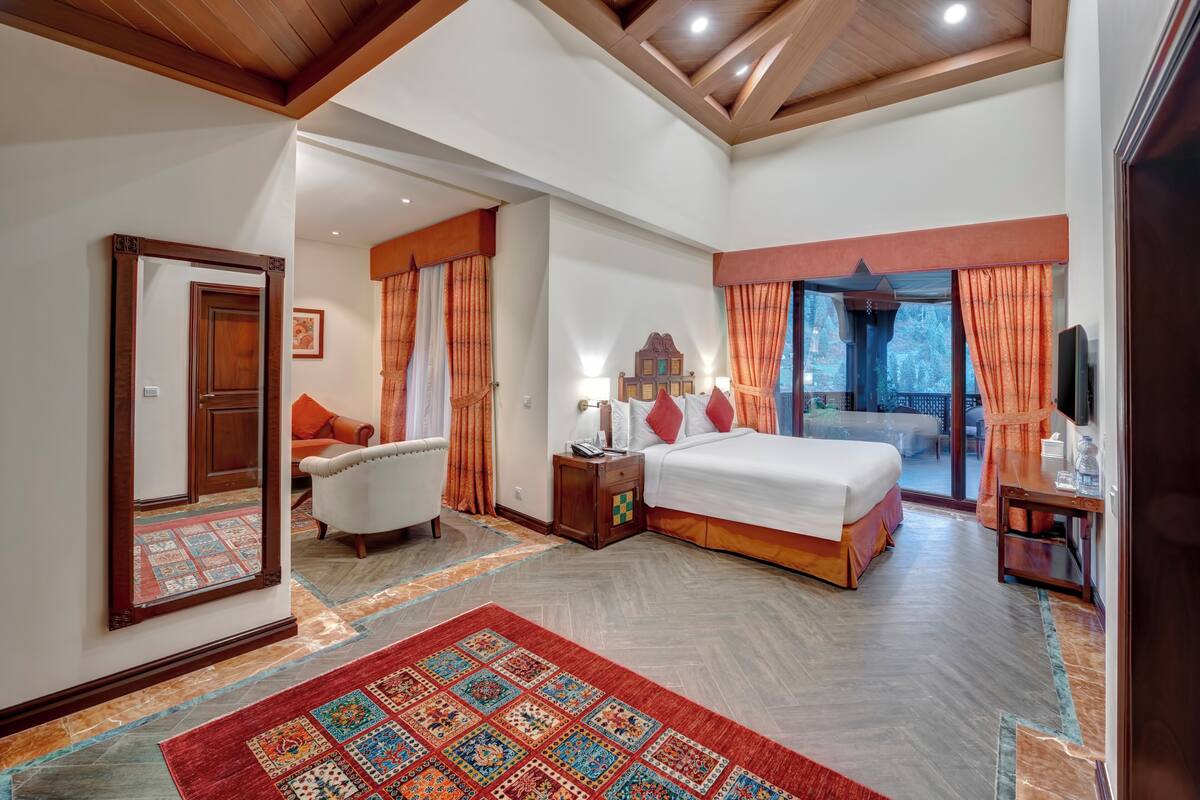 A spacious bedroom with a large bed, a sitting area with a chair, a nightstand, and a view of the outdoors.