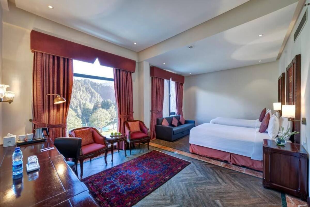 A hotel room with a large bed, a desk with a chair, a sofa, and a view of a mountainous landscape.