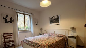 A bedroom with a bed, a chair, a nightstand, a lamp, a window with curtains, and a wall-mounted picture.