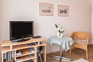 Une pièce avec un téléviseur à écran plat posé sur un meuble en bois, une chaise en osier, une petite table sur laquelle se trouve un vase de fleurs, et des tableaux encadrés accrochés au mur.