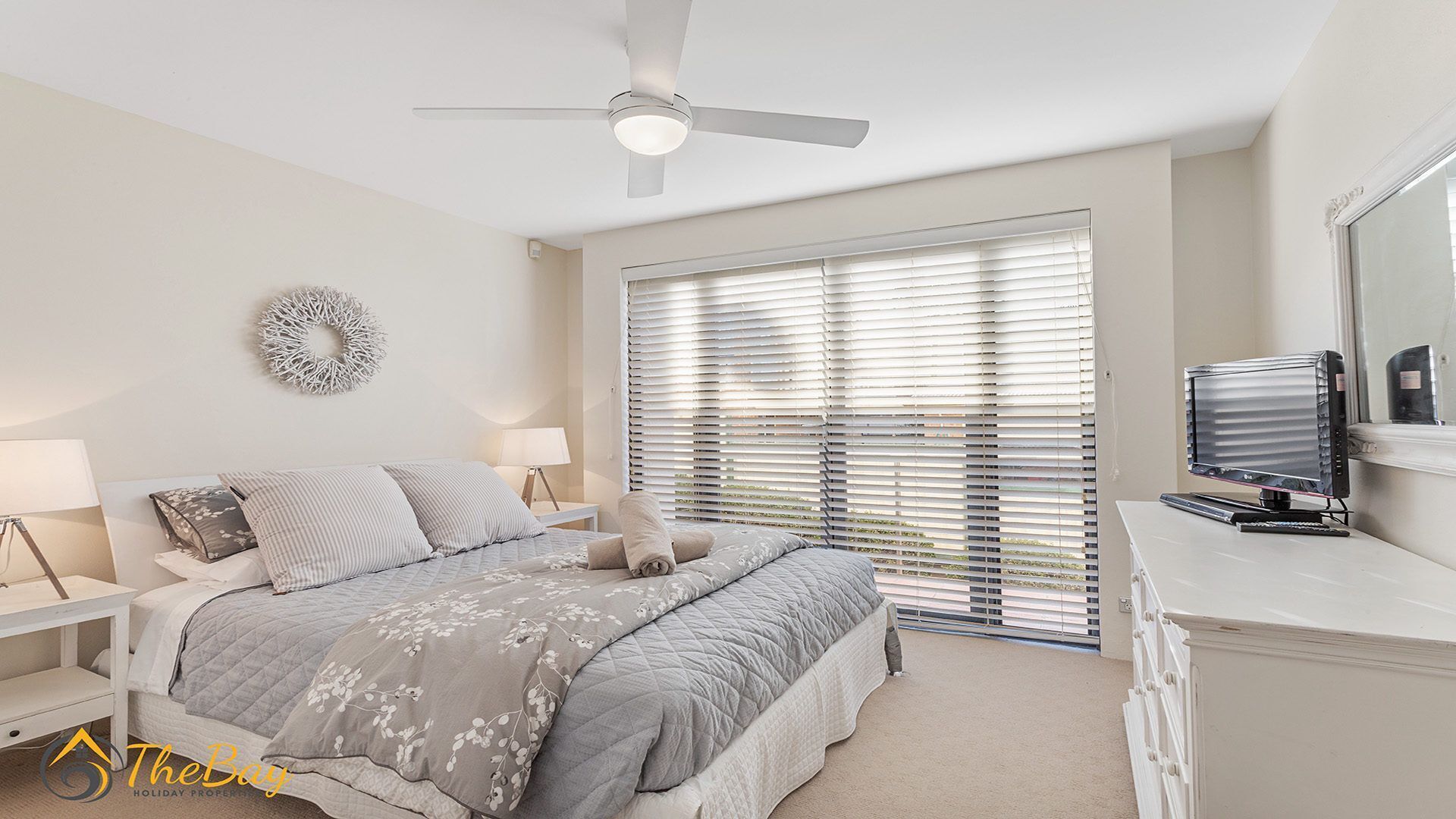 A bedroom with a bed, bedside tables, a ceiling fan, a TV, and a large window with blinds.