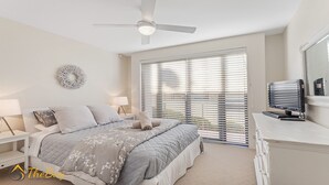 Un dormitorio con cama, mesitas de noche, ventilador de techo, televisor y un amplio ventanal con persianas.