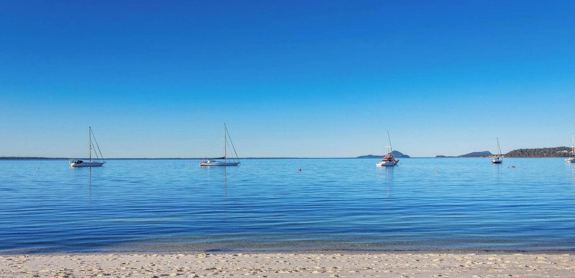 A beach with several sailboats anchored in calm waters, a clear sky, and a sandy shore.