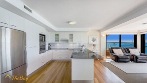Una cocina moderna con armarios blancos, electrodomésticos de acero inoxidable y una isla central. Hay un comedor con una mesa y sillas negras, y vistas al mar a través de amplias ventanas.