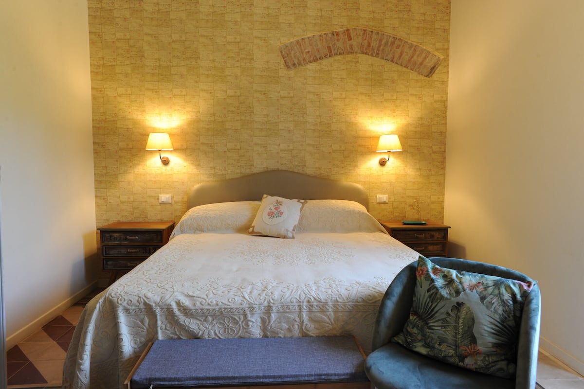 Ein Schlafzimmer mit einem großen Bett, zwei Nachttischlampen, einem Holznachttisch, einem Sessel mit einem blumigen Kissen und einem Wandbild.