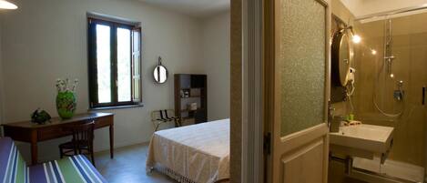 A bedroom with a bed, a desk, a chair, a window, and a bathroom with a shower and sink.