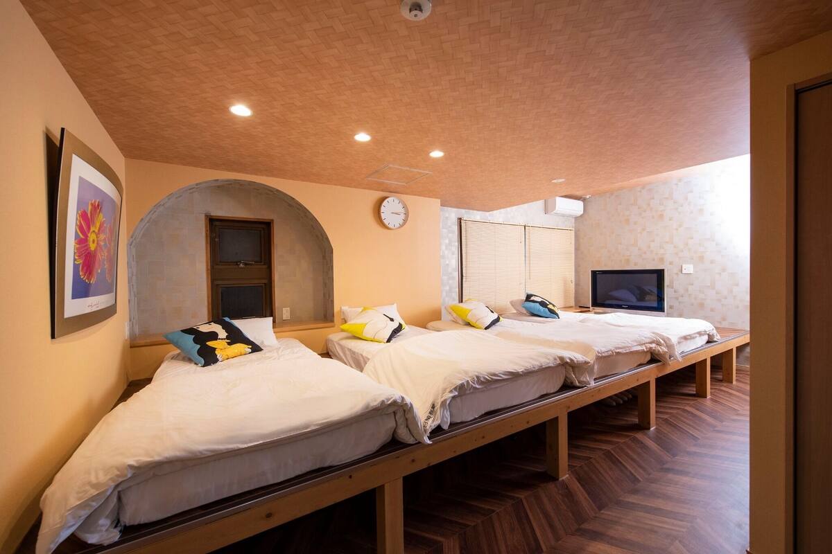 A hotel room with three single beds, a wooden bench, a TV, and a clock on the wall.