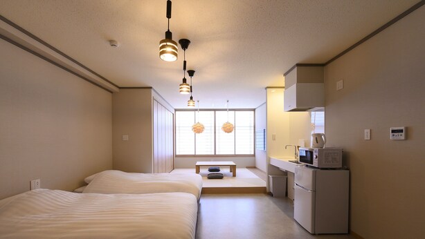 Una habitación de hotel compacta con dos camas, un kitchenette, una pequeña mesa y una ventana con persianas.