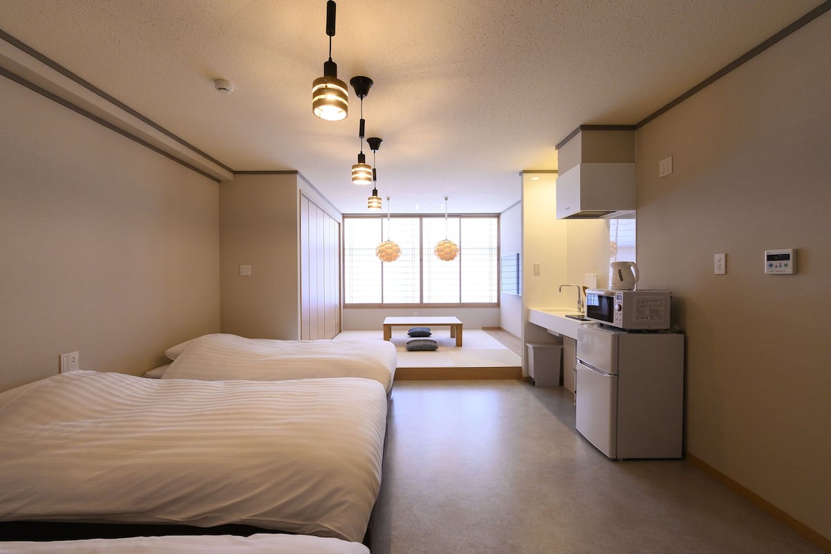 A compact hotel room with two beds, a kitchenette, a small table, and a window with blinds.