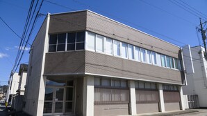 Un edificio de dos pisos con techo plano, ventanas amplias y entrada protegida por una reja.