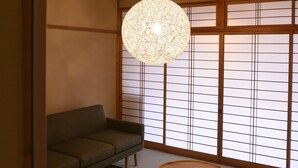 A traditional Japanese room with tatami flooring, a low wooden ceiling, a green sofa, two tatami mats, a round wooden table, and a hanging light fixture. - BEYOND HOTEL Takayama 4th (Takayama)