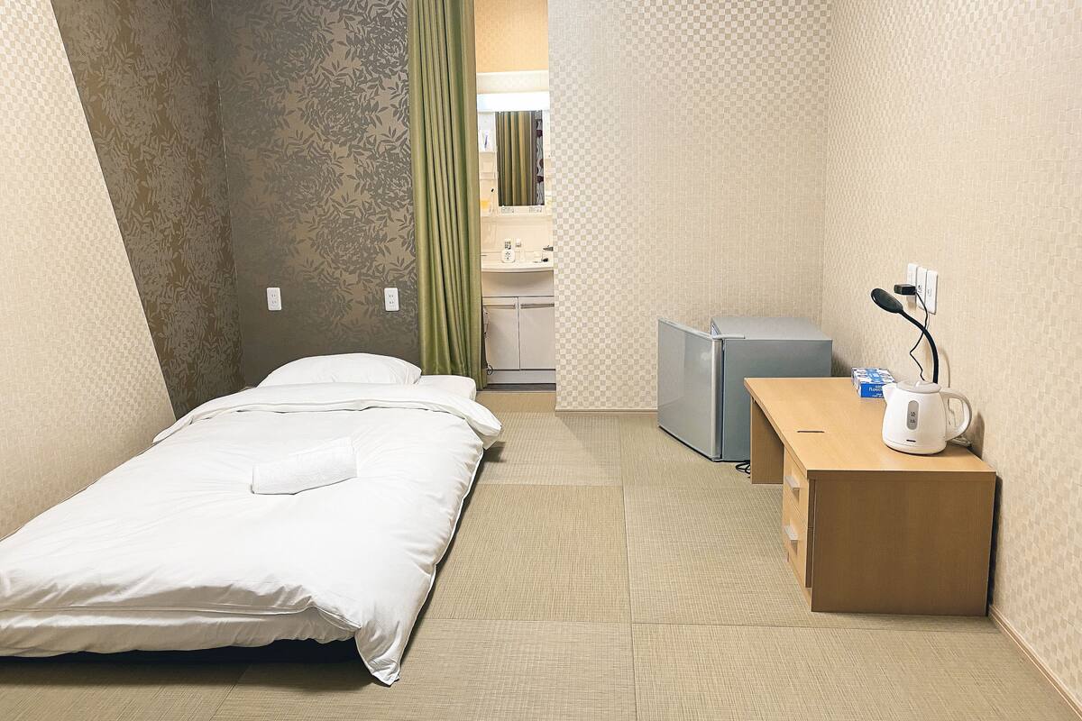A compact hotel room with a bed, a desk, a kettle, and a small refrigerator.