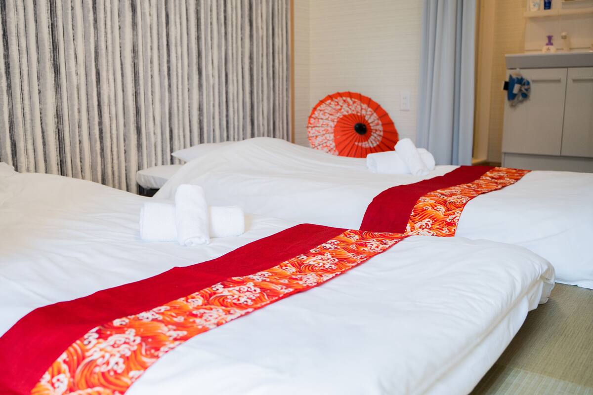 A hotel room with two beds, white linens, a red and orange patterned bed runner, and a red and white umbrella on the headboard.