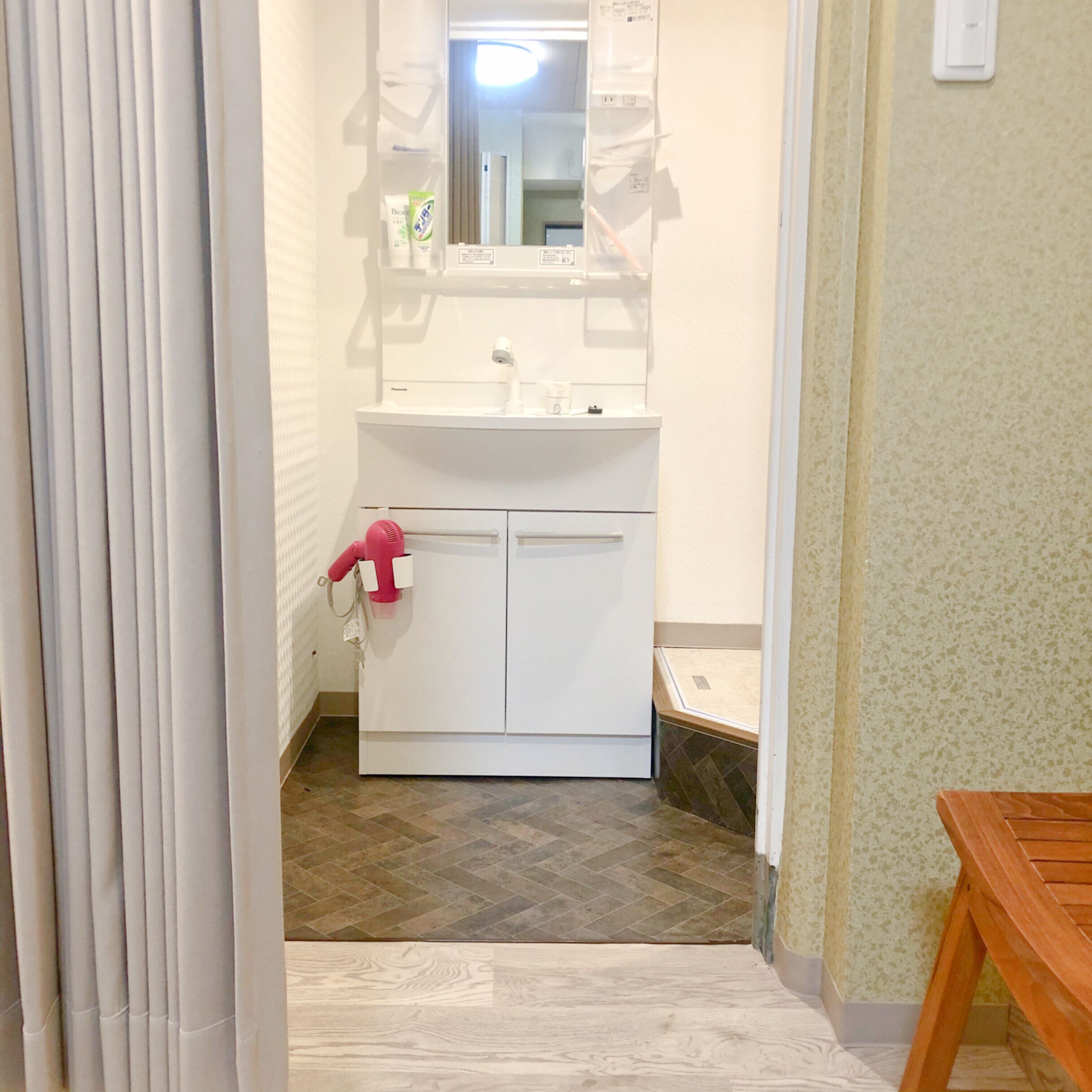 A bathroom with a white sink, mirror, and cabinets. There is a towel rack with a pink towel and a hairdryer. The floor is tiled, and there is a wooden bench in the corner.