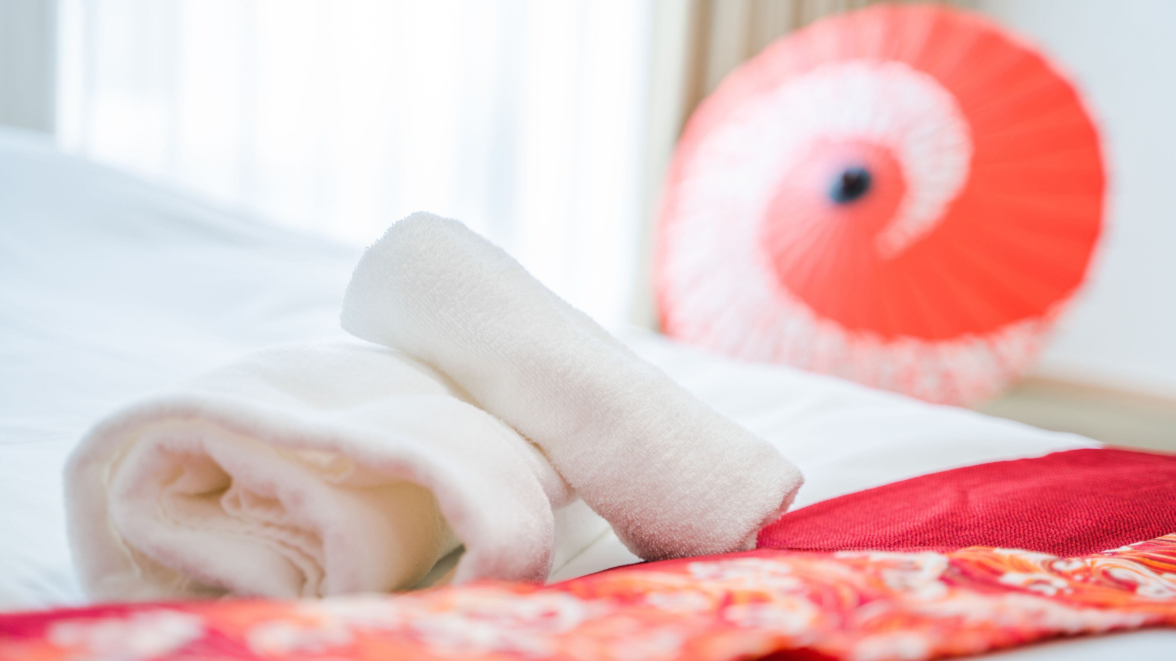 A neatly made bed with a rolled white towel and a red pillow.