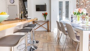 A modern kitchen with a wooden bar, a dining area with chairs, a TV mounted on the wall, and a brick accent wall. - Stroll To The City Centre In From Chic Refurbished Townhouse (Cardiff)