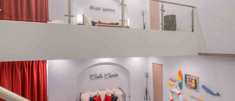 A hotel interior with a glass railing, a sofa with red and black cushions, and a wall with a surfing-themed poster.