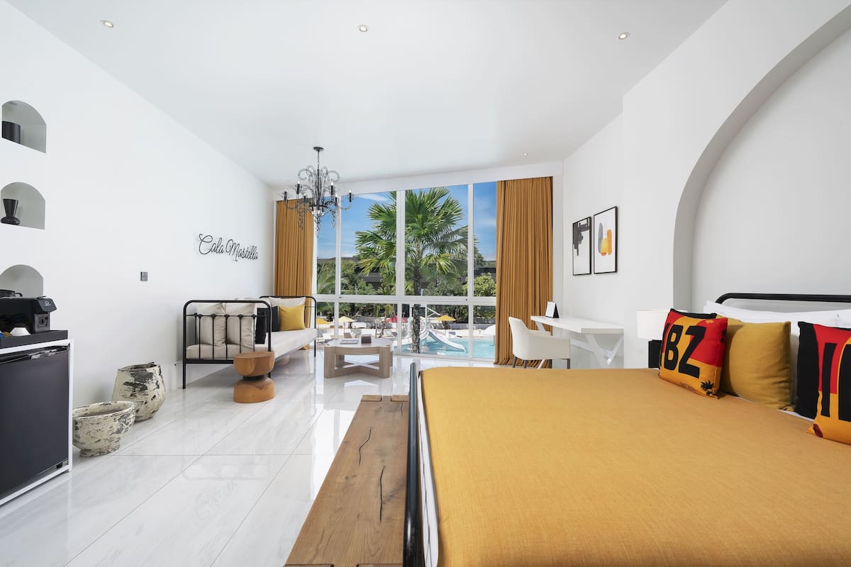 A modern hotel room with a large bed, a desk, a sofa, and a view of the pool.