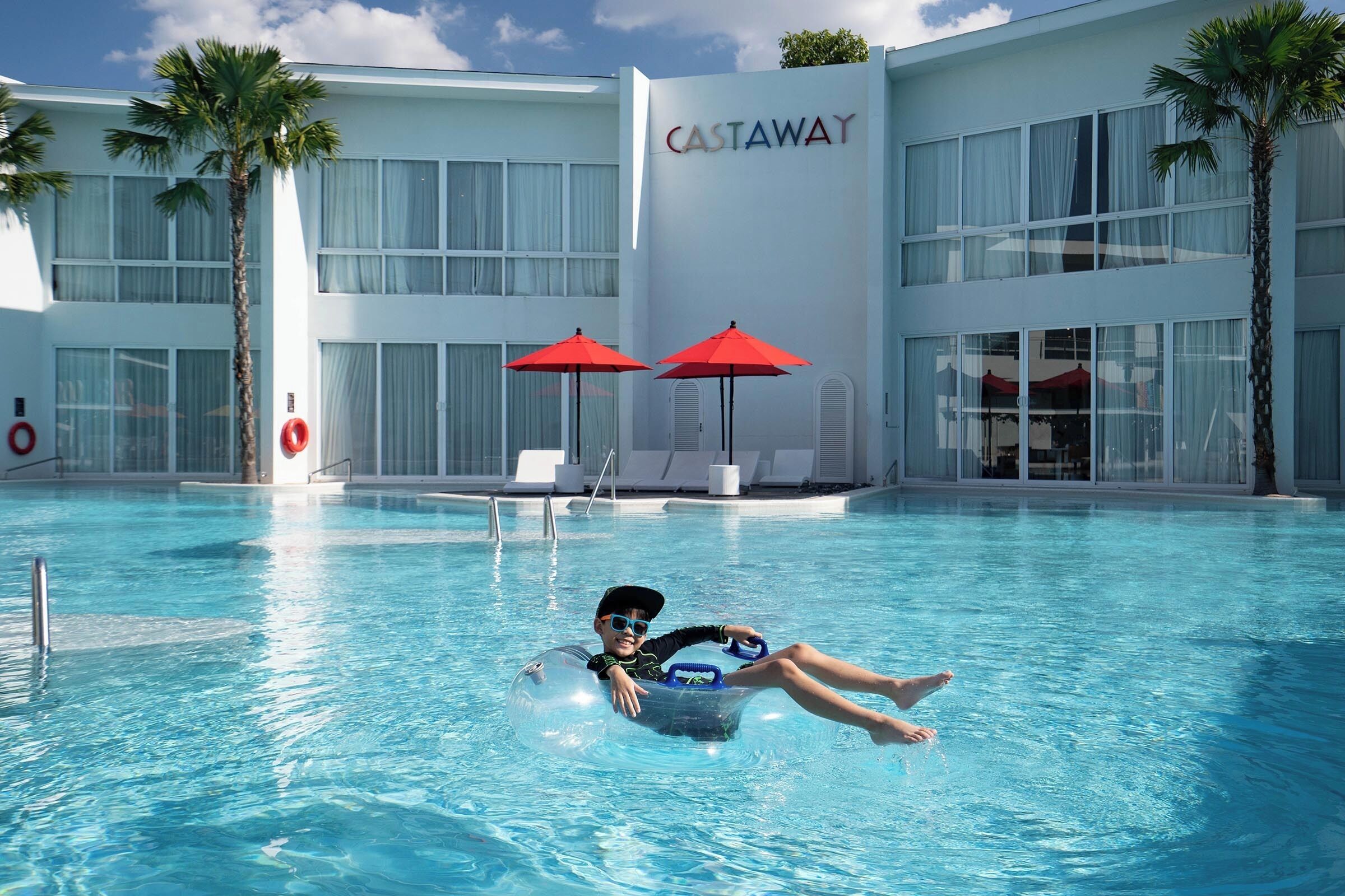 A person floating in a pool at a hotel with a large building in the background labeled 'CASTAWAY.'