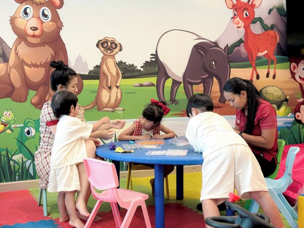 Niños participan en una actividad artística en una mesa con tapetes de juego coloridos y un mural de animales al fondo.
