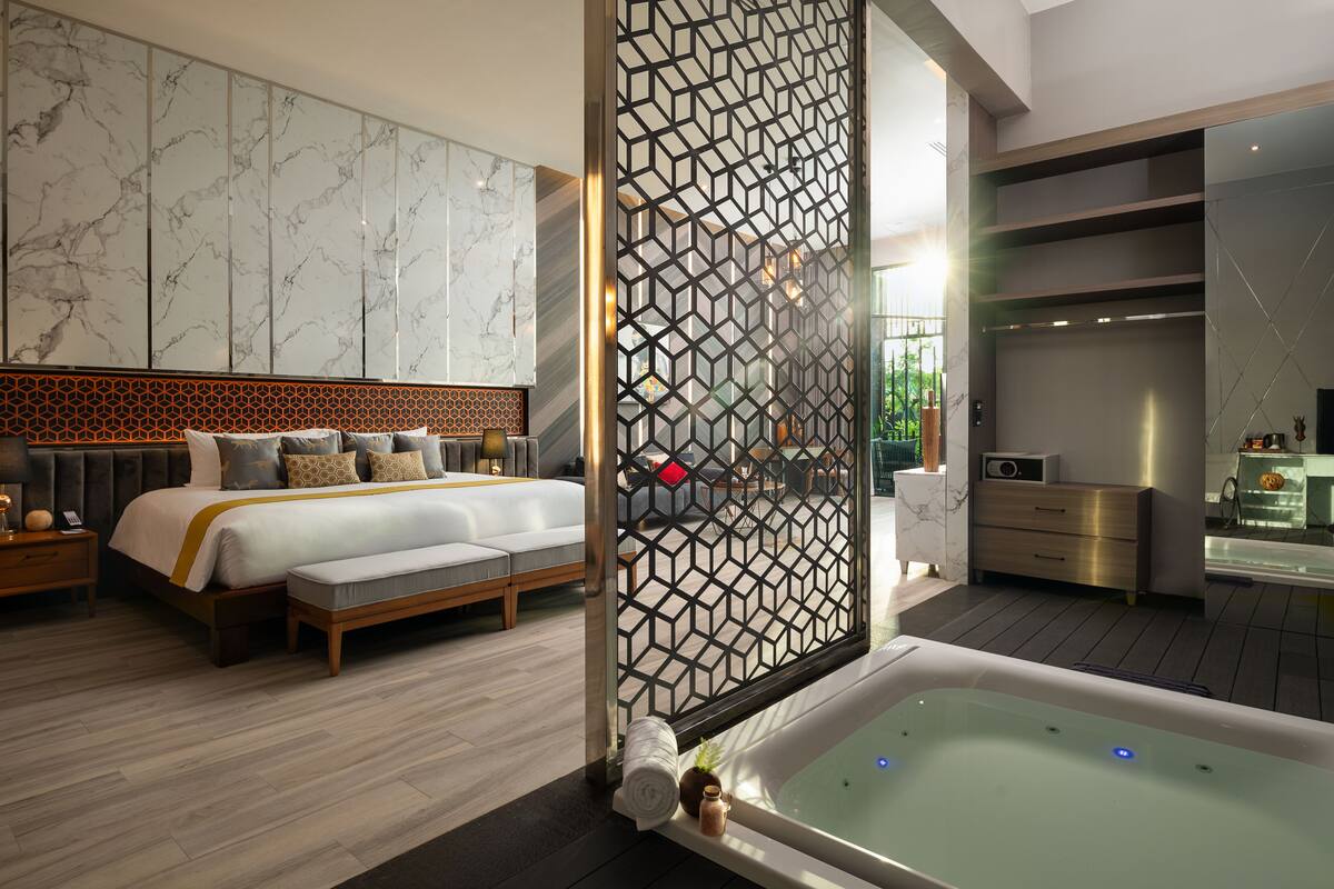 A modern bedroom with a large bed, a separate sitting area, a bathroom with a bathtub, and a decorative partition.
