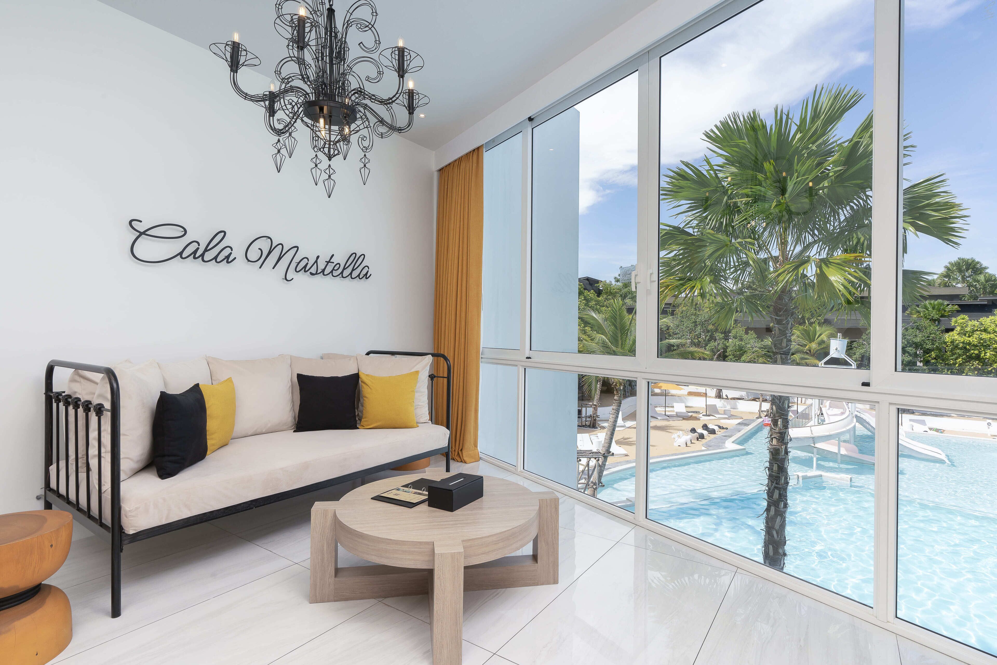 Ein modernes Hotelzimmer mit einer Couch, einem runden Couchtisch und einem großen Fenster mit Blick auf einen Swimmingpool und Palmen.