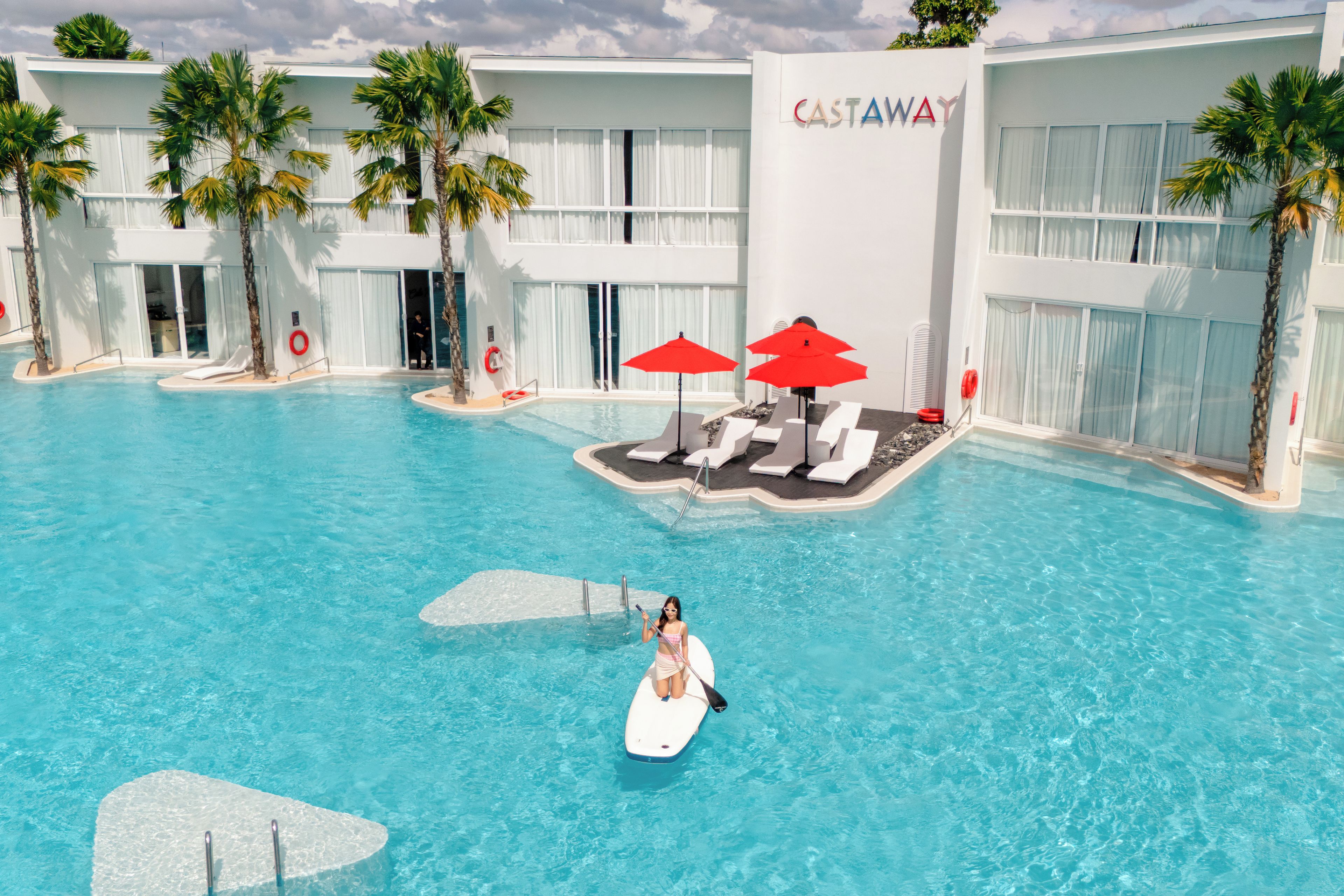 A swimming pool area with lounge chairs and umbrellas, surrounded by palm trees and a building with 'CASTAWAY' signage.