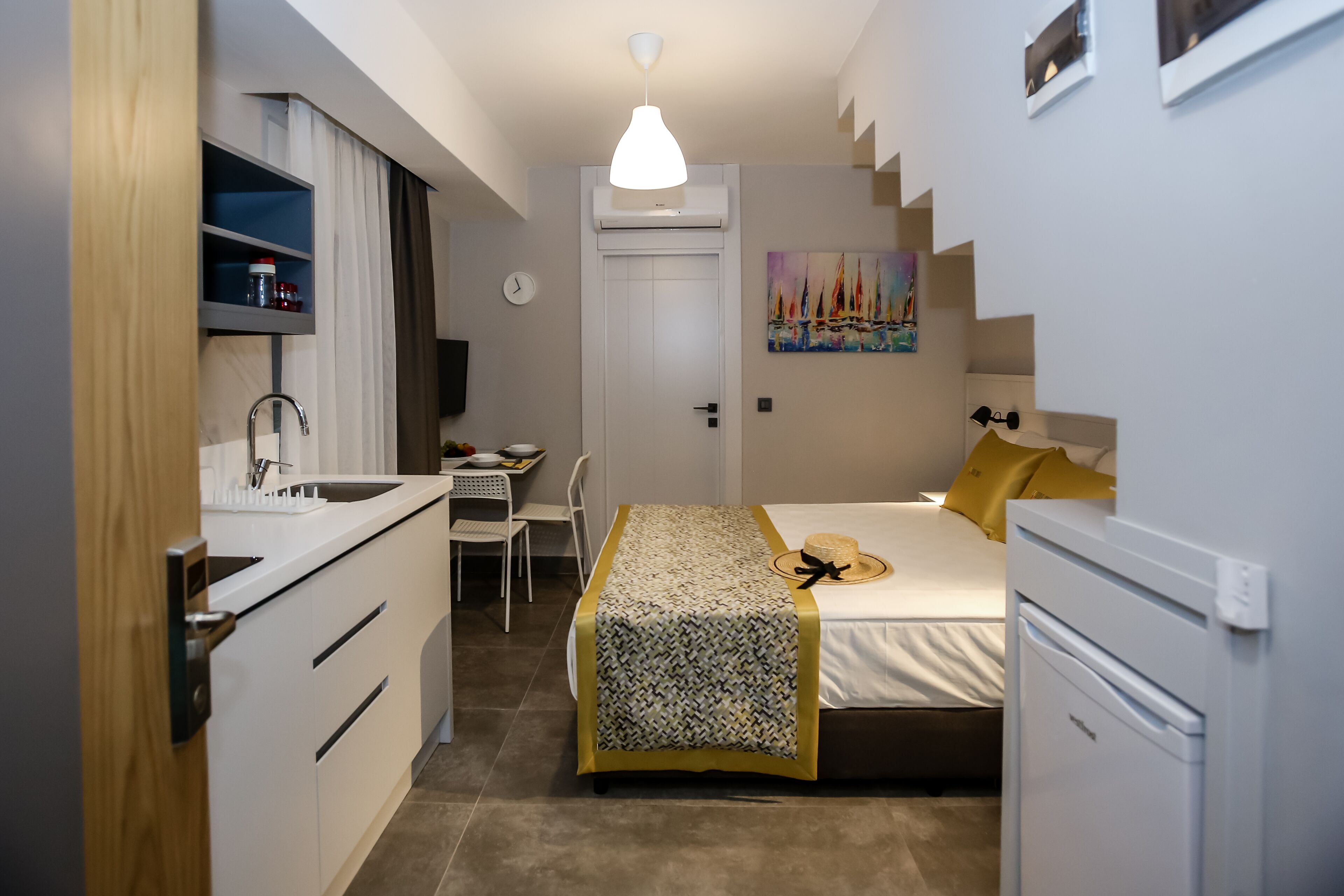 Ein kleines Hotelzimmer mit Küchenzeile, einem Bett mit gemustertem Überwurf, einem Schreibtisch mit Stuhl und einem Bild an der Wand.
