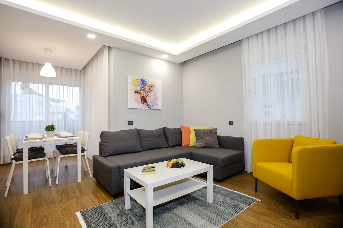 Un salon moderne avec un canapé gris, un fauteuil jaune, une table basse blanche et un coin repas comprenant une table et des chaises blanches.