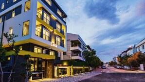A modern multi-story hotel with balconies, a well-lit entrance, and a paved street in the background.