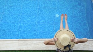 A person sitting by a pool with a straw hat, wearing patterned swimwear.