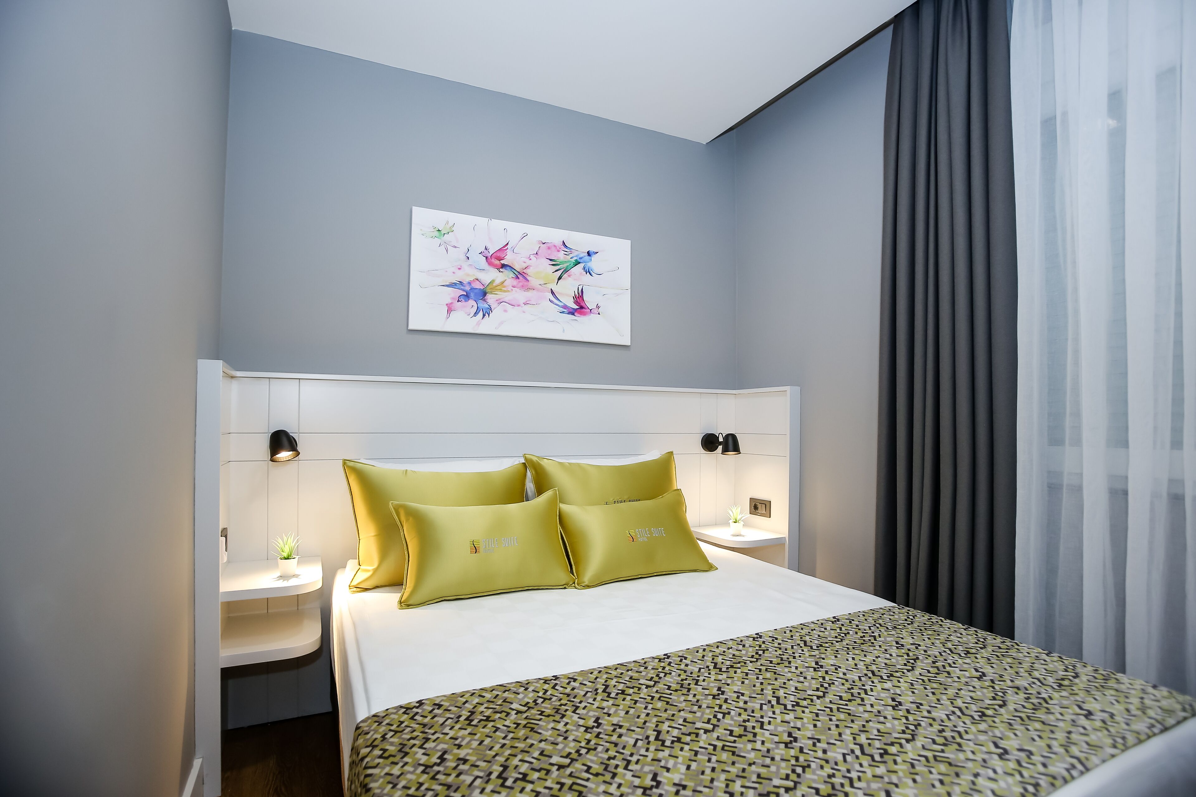 Une chambre d’hôtel avec un lit, deux tables de chevet, une lampe fixée au mur, une petite plante en pot et une fenêtre avec des rideaux.