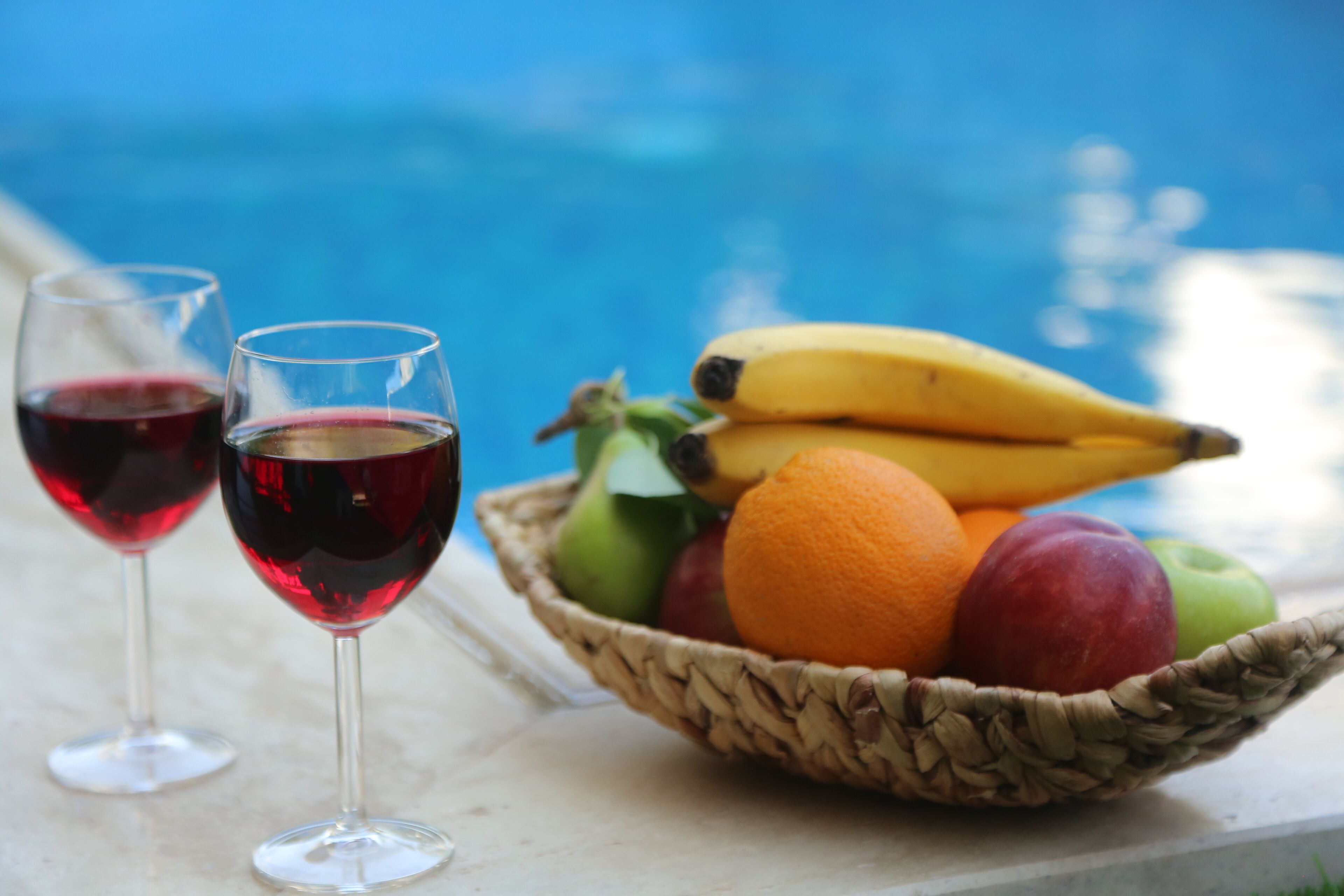 A poolside setting with two glasses of red wine and a basket of fruit including bananas, apples, and oranges.