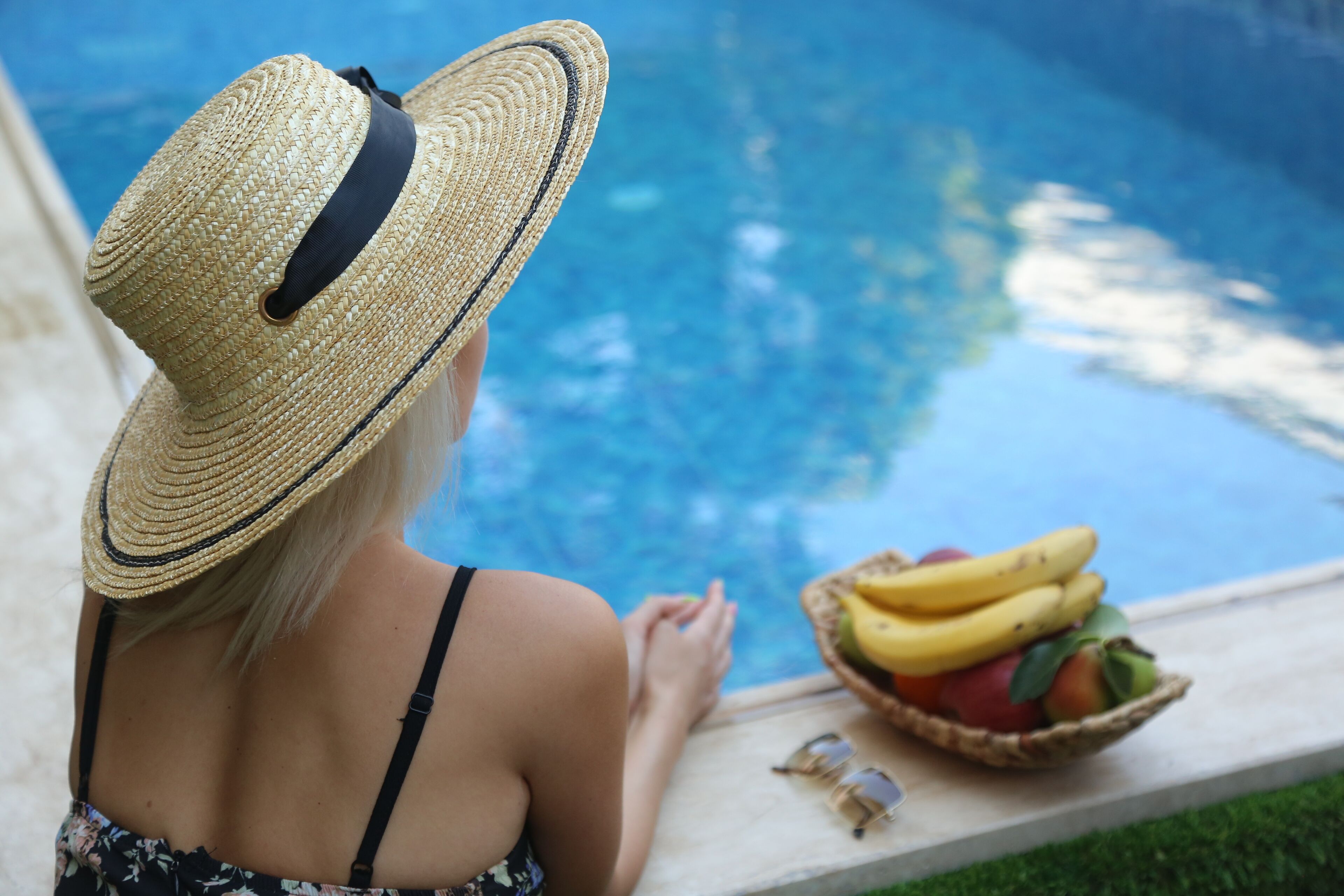 A person sitting by a pool with a basket of fruit and sunglasses on the edge.