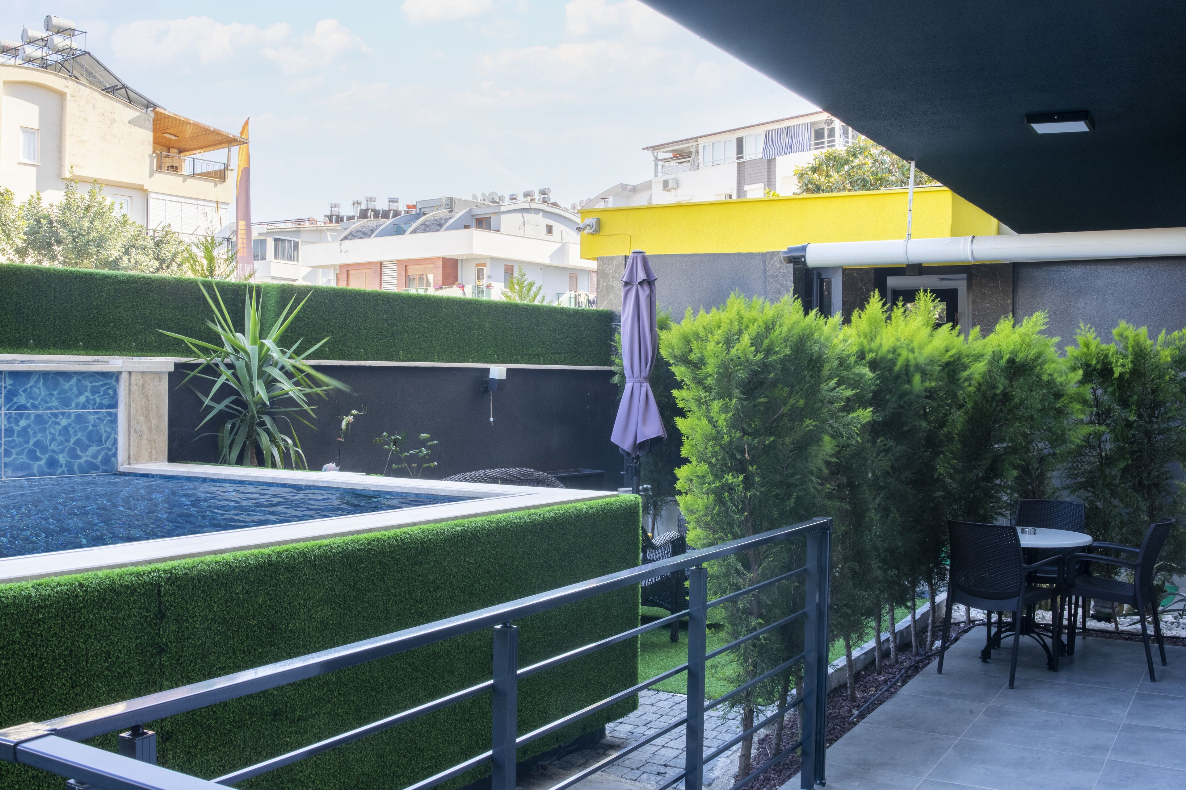A rooftop pool area with a clear view of the surrounding buildings, a seating area with chairs, and a pergola with a purple umbrella.