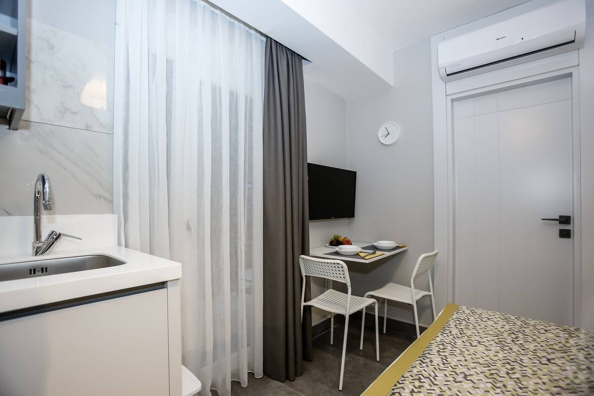 Une chambre d’hôtel compacte avec un lavabo blanc, une petite table avec une chaise, un téléviseur à écran plat et une porte équipée d’un appareil de climatisation.