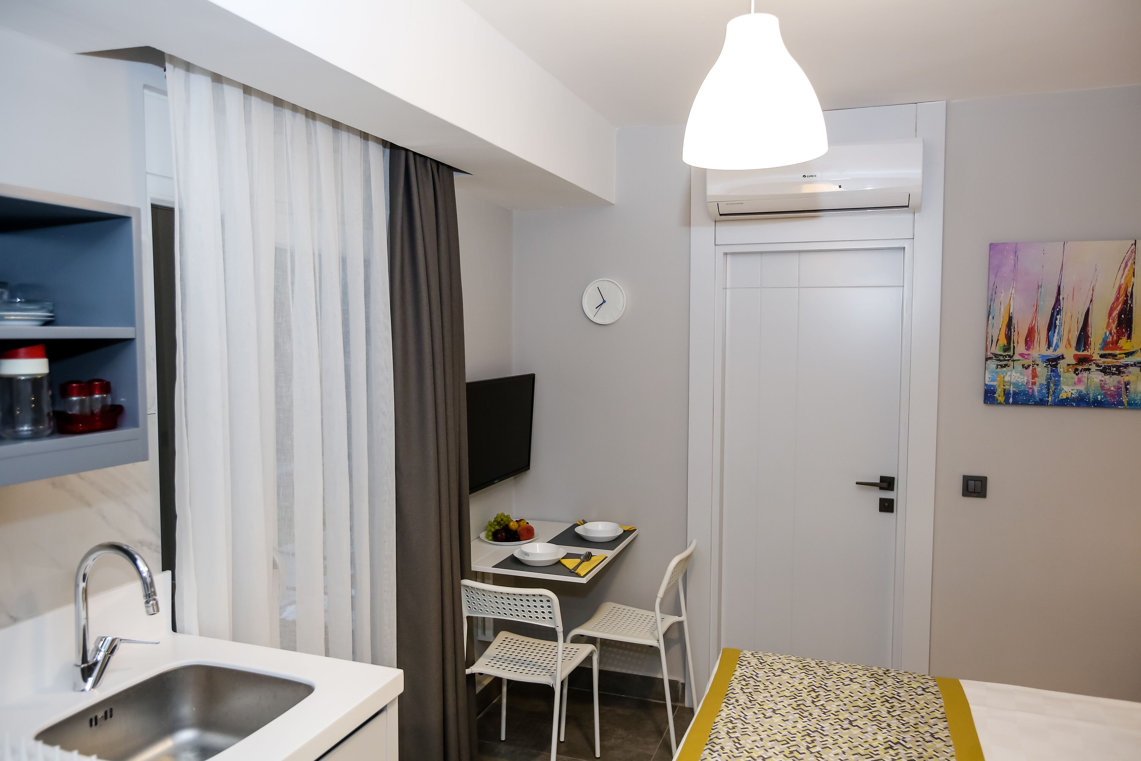 Camera d'albergo compatta con angolo cottura, zona pranzo con tavolo e sedie, un letto, TV a parete e un quadro con dei velieri.