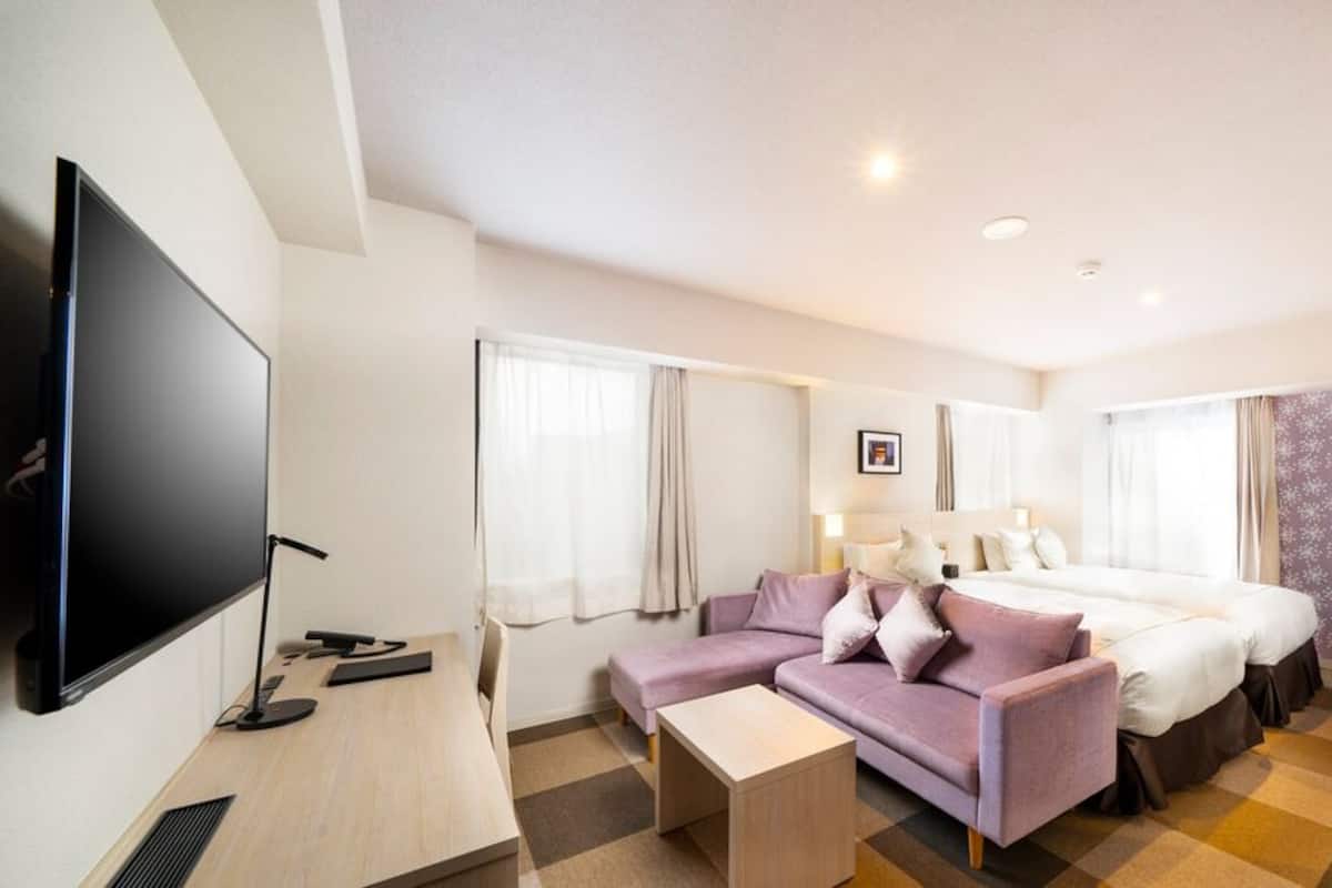 A hotel room with a flat-screen TV, a sofa, two beds, a desk, and a patterned wall.