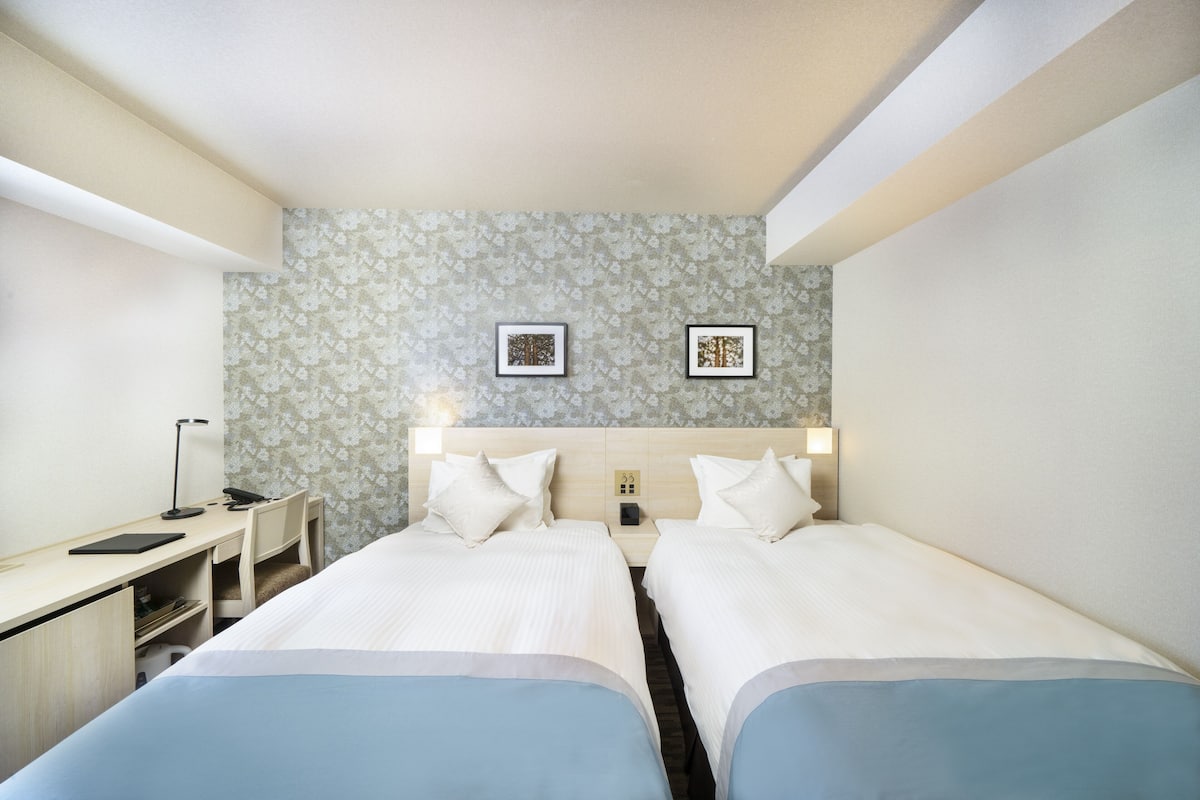 A hotel room with two single beds, a desk with a chair, a nightstand, and two framed pictures on the wall.