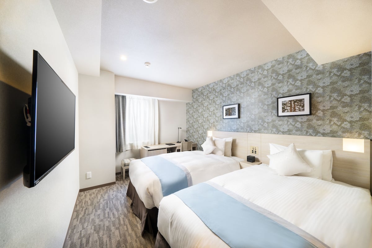 A hotel room with two beds, a desk, a chair, a TV, and a patterned wall.