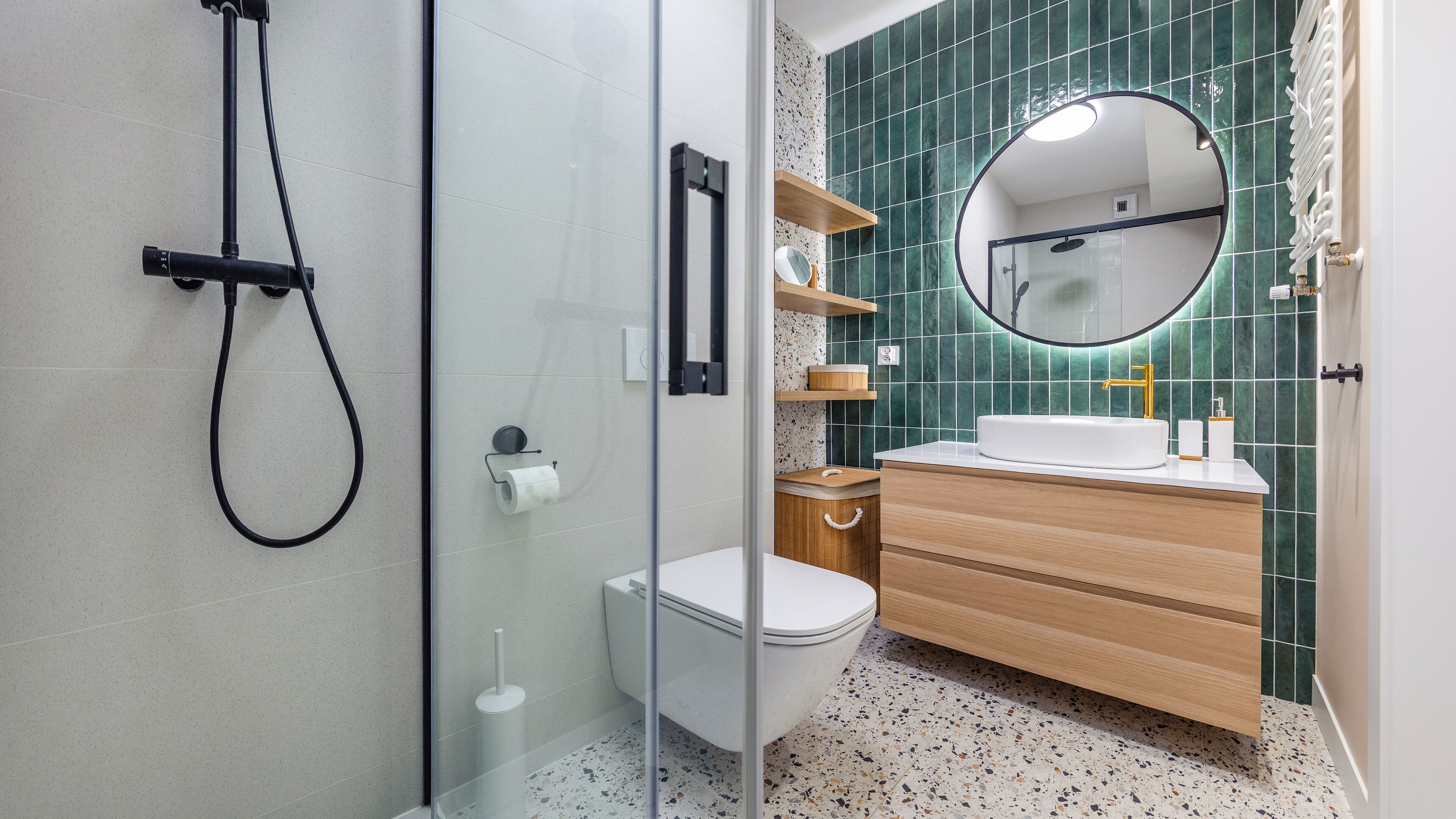 Un baño moderno con cabina de ducha de vidrio, inodoro blanco, tocador de madera con lavamanos blanco y salpicadero de azulejos verdes.