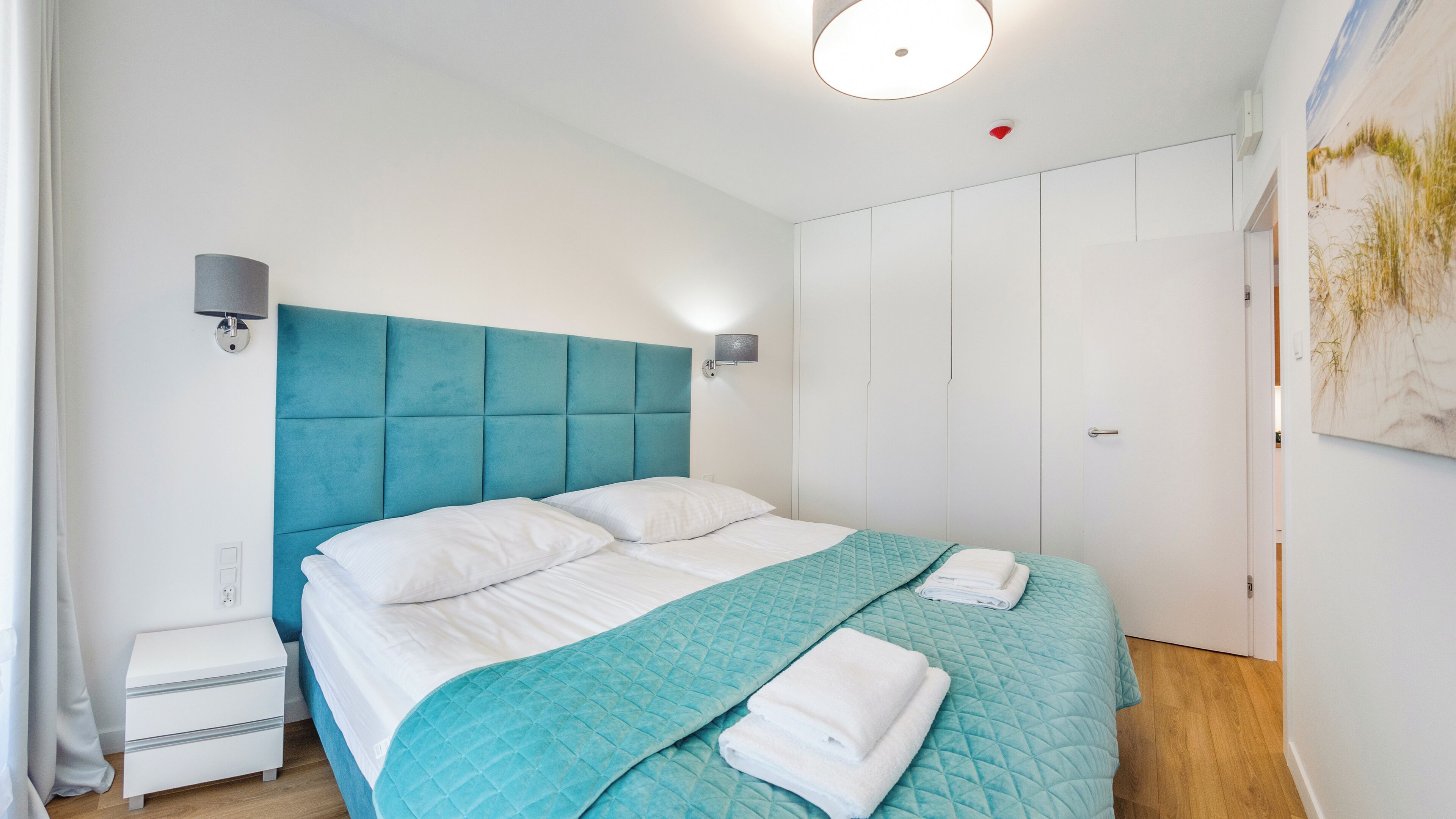 A bedroom with a teal headboard, white bedding, a bedside lamp, a nightstand, and a door with a painting.