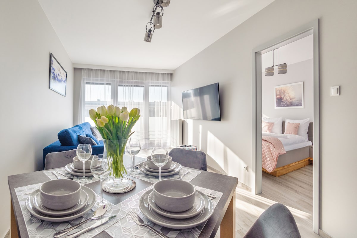 A modern living room with a dining area set for four, a blue sofa, a flat-screen TV, and a large window with sheer curtains.