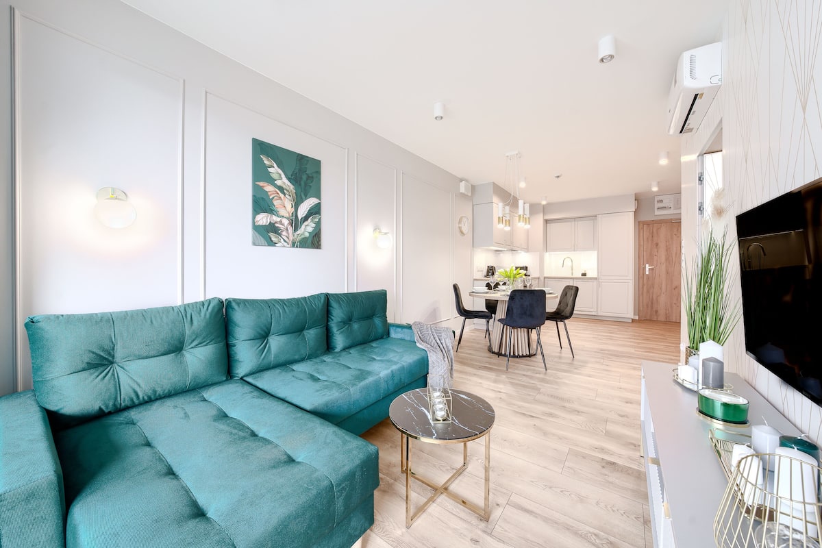 A modern living room with a teal sofa, a small round table, and a dining area in the background.